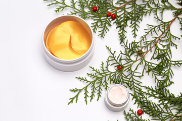 green Christmas tree branches, red new year berries, cosmetic products in jars, eye patches, face cream on a white background, top view, flat lay, place for text