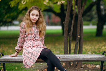 Obraz premium A cute teenage girl with lush hair in a beautiful plaid dress sits on a park bench