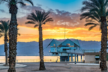 Colorful sunrise above mountains and public beach with promenade and decorative palms as foreground, Red Sea, Middle East © sergei_fish13