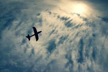 Silhouette of toy airplane flying  to the sun on the cloudy blue sky background. Children's plane in the air.