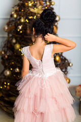 Little girl in a chic pink dress stands with her back to the camera and looks at gifts under the tree