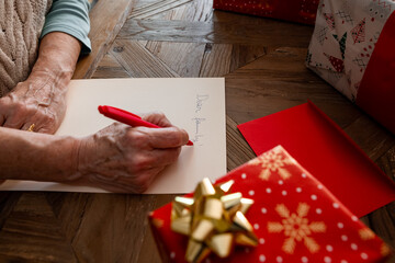 Close up of senior woman's hand writing a letter on yellow paper on wooden table, to her family for Christmas, with wrapped gifts on a side. Social distance, self isolation, quarantine concept.
