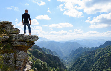 Naklejka na ściany i meble A young brutal man stands on the edge of a mountain cliff and meditates, enjoying the view of mountain peaks.