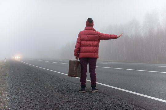 A young woman in a red jacket with a suitcase in her hands stands on the side of the road, raises her hand and asks passing cars to stop. Fog on the roads. Hitching a ride.