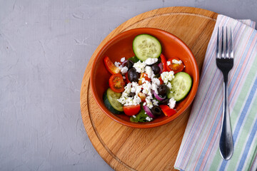 A brown plate of greek salad on a round stand next to a napkin and a fork on a gray concrete background. Copy space.