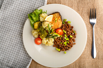 Baked fish with broccoli and lentils in a white plate with rosemary and lemon on a napkin and a round stand next to a fork.