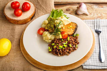 Baked fish with broccoli and lentils in a white plate with rosemary and lemon on a napkin and a round stand next to a fork and vegetables.