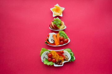 Plate in the form of a Christmas tree with vegetables, fruits, mushrooms and berries. Red background. The concept of vegetarian treats for the holiday. Copy space