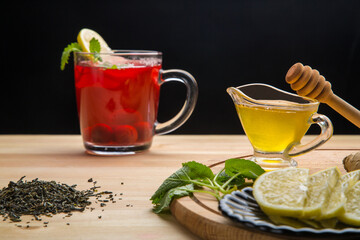 Berry tea in a cup next to lemon, honey, lemon balm and a teapot with a drink on the table.