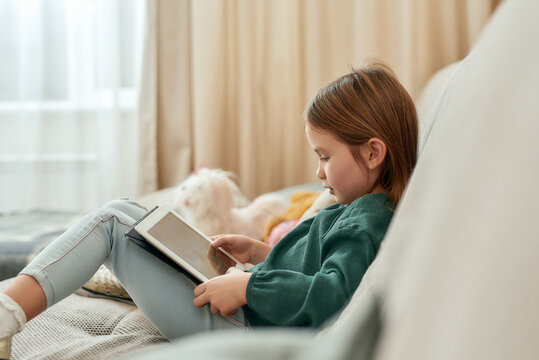 A cute small girl playing videogames on a tablet while sitting on a sofa