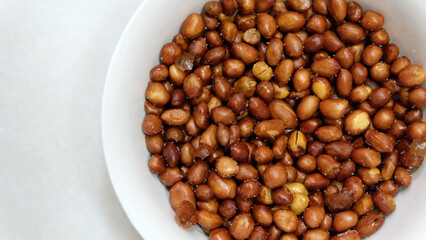 A bowl of fried peanuts with skin, lightly salted, in a white bowl. Top view.
