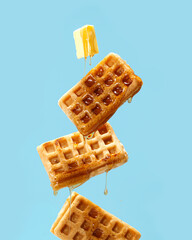 Flying waffles and butter getting dripped with maple syrup over a light blue background