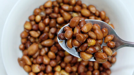 A spoonful of fried peanuts, coated with skin. With a bowl of peanuts in the blurred background.