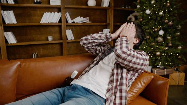 Depressed young man drinking alcohol at home. Unemployed male, drunk and continues drink wine straight bottle on couch near Christmas tree. Addiction, loneliness, nervous breakdown new year holiday