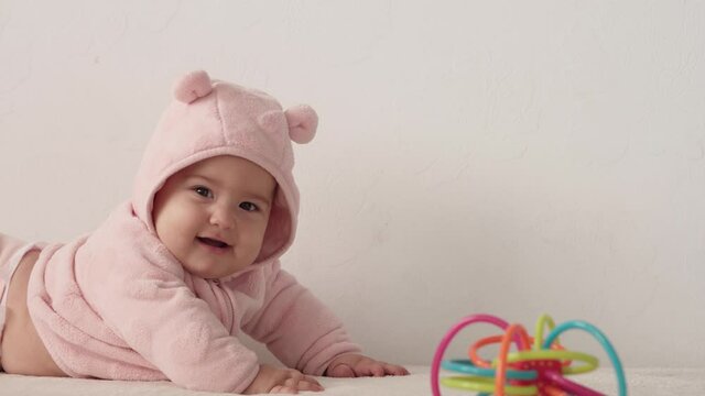 infant, childhood concept - close-up of smiling happy plump funny brown-eyed chubby face of newborn kid awake. toothless 7 month baby grimaces, lie on soft bed in pink with teether at home