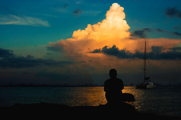 Silhouette of a man sitting in the sea in sunset and a tourism ship.   A beauty sky clouds colorfull of light of the sun.