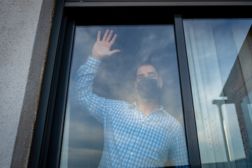 Bearded man wearing blue shirt and mask to prevent coronavirus looking out the windows