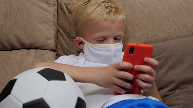 A Little Boy In A Medical Mask Is Lying On The Couch At Home With A Soccer Ball And Playing Games On His Phone. Quarantine And Distance Learning Due To The Coronavirus Outbreak.