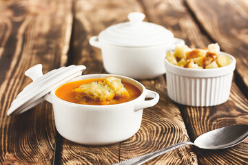 Vegetarian spicy autumn pumpkin cream soup served in white cocotte with croutons and pumpkin seeds on wooden table. Seasonal autumn vegetarian food.