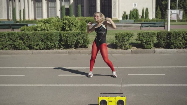 Wide shot of confident slim woman doing gymnastics or aerobic in 1980s on city road. Portrait of happy young Caucasian woman dancing and exercising to disco music from yellow tape recorder.