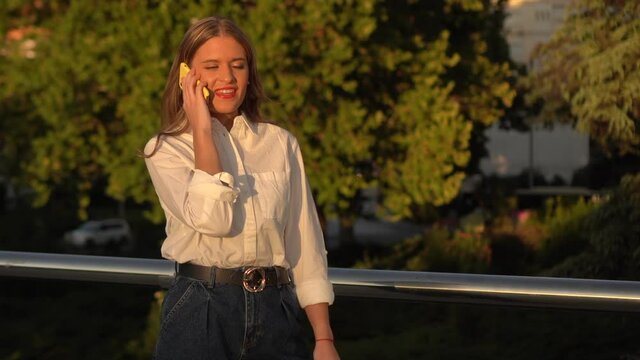 A beautiful young woman with long blonde hair talking on her cell phone on the street while standing. She is wearing a white shirt and blue jeans.