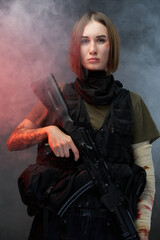 Seductive and tattooed woman dressed in modern and military body armour holding assault rifle in dark foggy background.