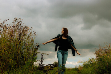young girl in a denim jacket, jeans, standing in a field dancing on a background of cloudy sky