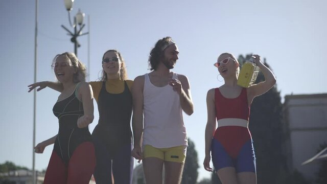 Group of positive 1980s friends in colorful sportswear and sunglasses walking in sunlight on urban street listening to disco music from vintage yellow tape recorder. Happy Caucasian people in 80s.