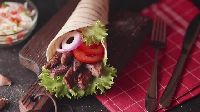 Handmade wrap shawarma is lying on the wooden cutting board. 
Shawarma is made with tortilla, beef, tomato, onion and lettuce on a black table.