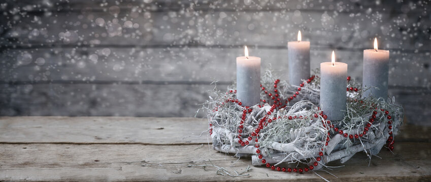Advent wreath of white painted branches with burning candles and a red chain, tradition in the time before Christmas, rustic wooden background with snowy bokeh and copy space, panoramic format