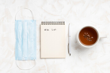 Christmas wish list in notepad with pen,mask and tea on white wooden bacckground