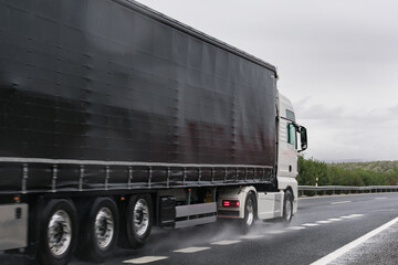 Truck with a black semi-trailer driving on the highway on a rainy day.