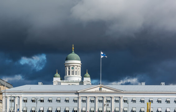 Main Green Dome Of White Helsinki Cathedral And Top Of Helsinki City Hall With The Flag Of Finland In The Roof Against Dark Stormy Sky.