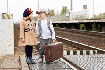 A little boy and a girl are sad on the platform near the railway tracks. Dressed in old clothes. Girl sitting on an old suitcase