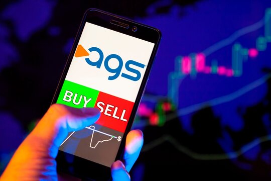 Company logo PlayAGS Inc on smartphone screen, hand of trader holding mobile phone showing BUY or SELL on background of stock chart. Russia, Stavropol - May 29, 2019