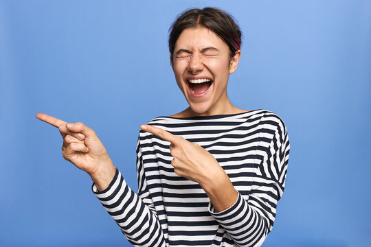 Isolated shot of emotional overjoyed young Caucasian woman in striped shirt posing with eyes closed and mouth opened, screaming, pointing fore fingers sideways, excited with big sale prices