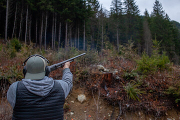 A mean wearing a vest, hat and ear protection holds an old 12 gauge shotgun, aiming and shooting at orange clay pigeons to practice. Pump action shotgun with a wooden stock.