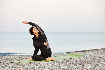 Young sporty woman with short hair in black sportswear practices yoga, relaxation and meditation on mat by the sea, Ardha Matsyendrasana, nature background, horizontal