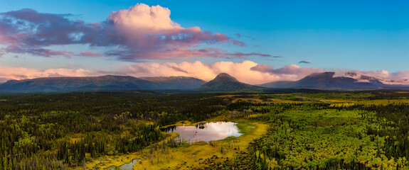 Naklejka na ściany i meble Peaceful Panoramic View of Pond and Marshland, surrounded by Forest and Mountains in Canadian Nature. Sunset Sky. Aerial Drone Shot. West of Whitehorse, East of Haines Junction, Yukon, Canada.