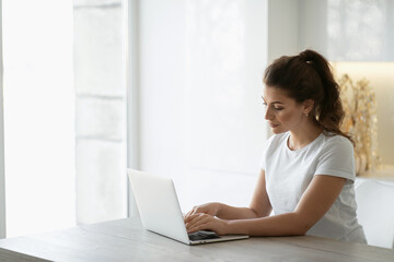 student Manager prints a message and writes a distance learning paper on a computer or laptop. A young woman is sitting at home in the kitchen in front of a gadget, looking at the monitor