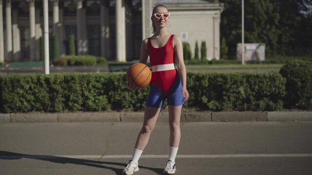 Wide shot portrait of confident smiling Caucasian woman in retro sportswear bouncing basketball ball outdoors and looking at camera. Slim beautiful 1980s sportswoman posing on city street in sunlight.