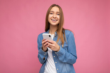 Image of a beautiful shocked young blonde woman posing isolated over pink wall background using mobile phone.