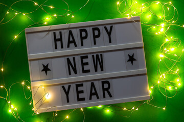 White letter lightbox with greeting Happy New Year and garland on green background. Top view