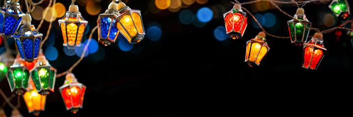 Fairy christmas lights with vintage lantern shaped lamps glowing on black bokeh background, close view for web banner with copyspace. Colorful garlands of Xmas and New Year winter holiday celebration 