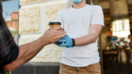 Close up of hands of man receiving hot coffee drink from cafe assistant in protective gloves while collecting his takeaway order during coronavirus lockdown