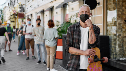 Portrait of middle aged man wearing mask holding longboard, looking at camera while waiting to collect his takeaway order from the pickup point during lockdown