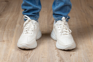 Standing feet shod in white lace-up chunky sole sneakers and blue jeans on the brown floor. Pair of new comfortable shoes for active lifestyle, everyday life and sports.