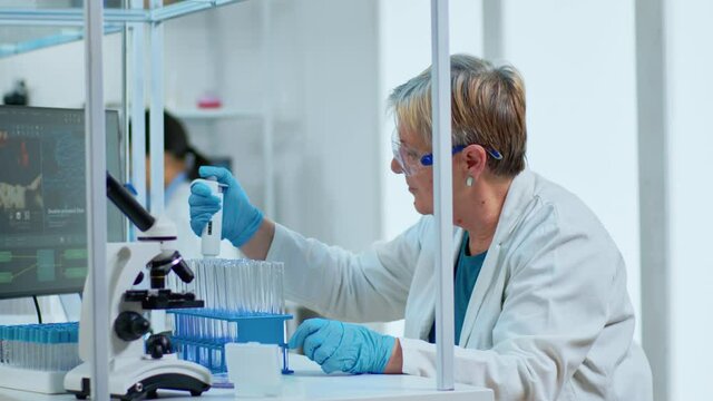 Senior scientist woman pipetting liquid to test tube in modern equipped lab. Multiethnic team examining virus evolution using high tech for scientific analysis of treatment development against covid19