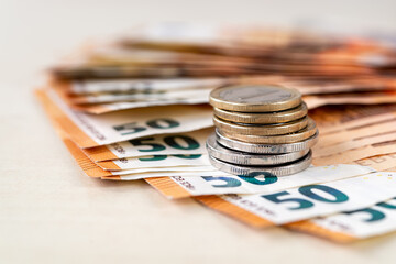 Stack of coins on 50 euro banknotes folded in a fan on a beige tabletop. Wealth, cash savings, accounting of income and expenses concepts. Shallow depth of field.