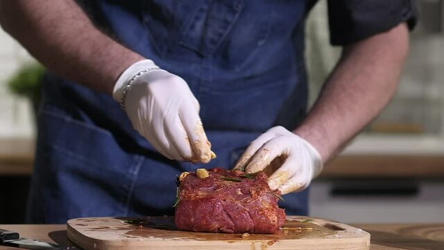 Preparing food in home kitchen. gloved cook spreads spices rosemary garlic pepper on large piece of meat and mixes it for baking in oven or cooking meat on  grill. Delicious dish for dinner